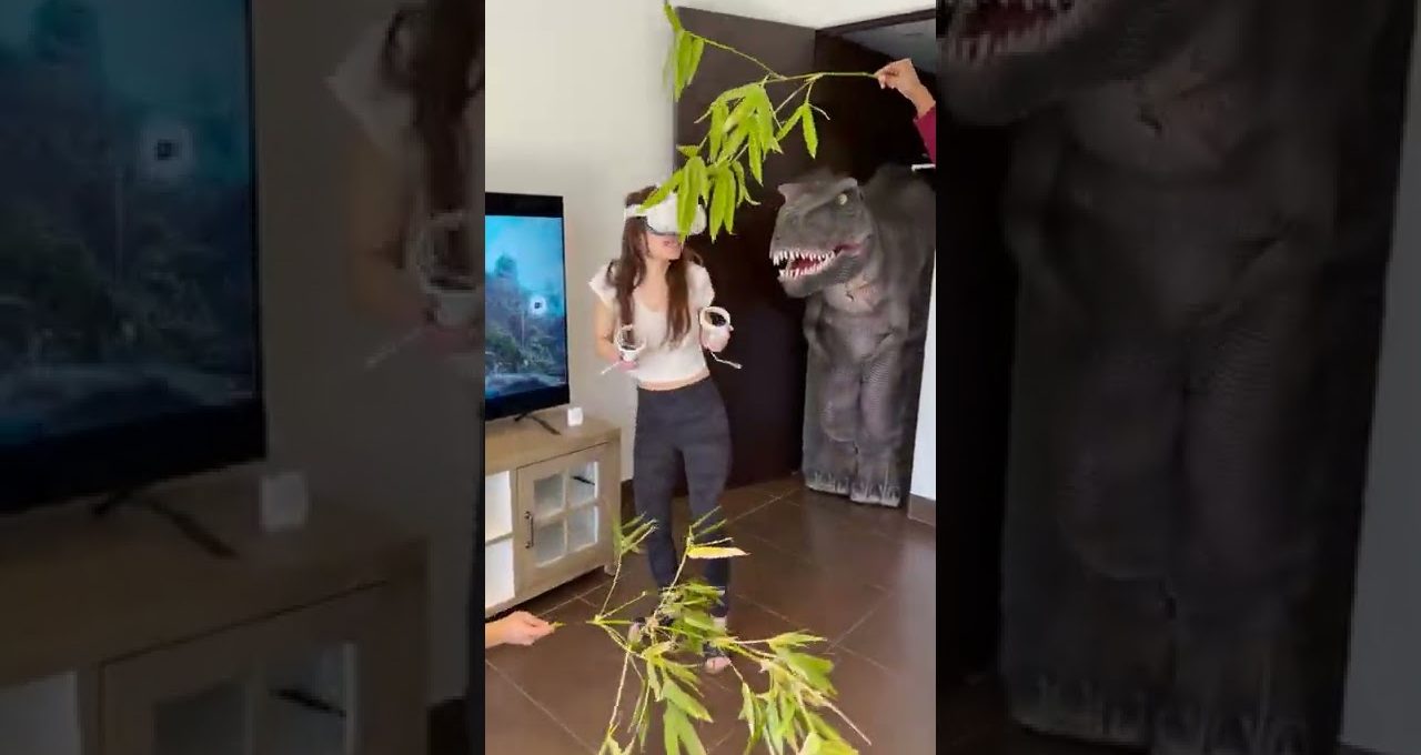 Woman wearing VR headset reacts as a person dangles branches beside a life-size dinosaur prop