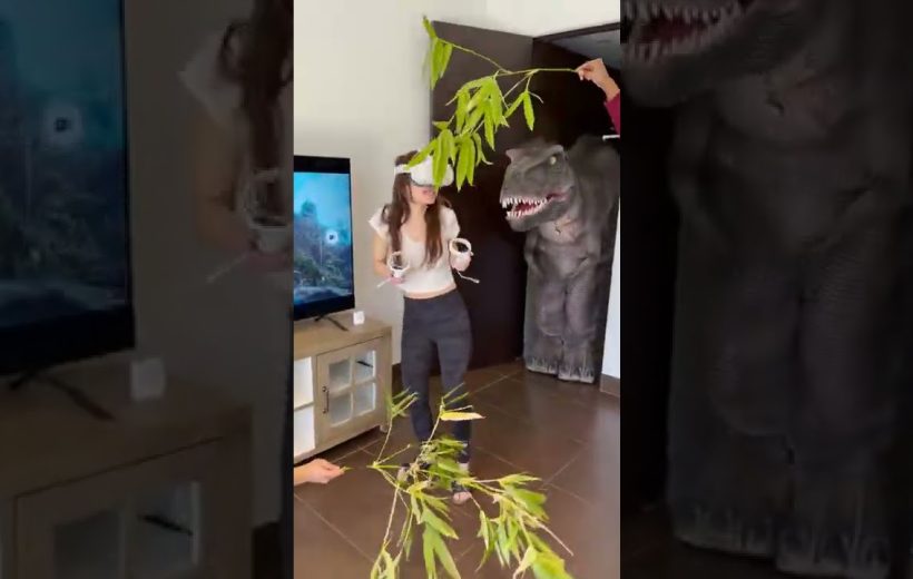 Woman wearing VR headset reacts as a person dangles branches beside a life-size dinosaur prop