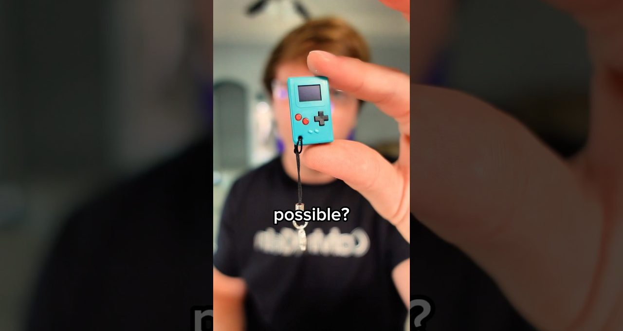 hand holding a tiny turquoise handheld game console charm close to camera, blurred person behind