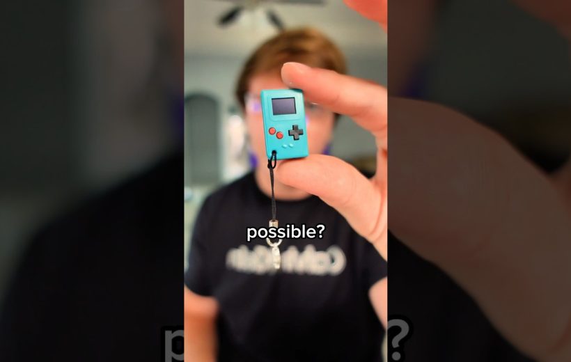 hand holding a tiny turquoise handheld game console charm close to camera, blurred person behind