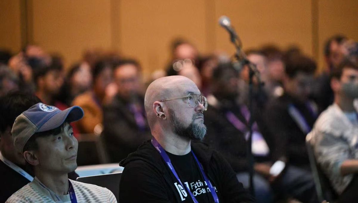 Bald man with glasses and lanyard attentively listening at a crowded conference session