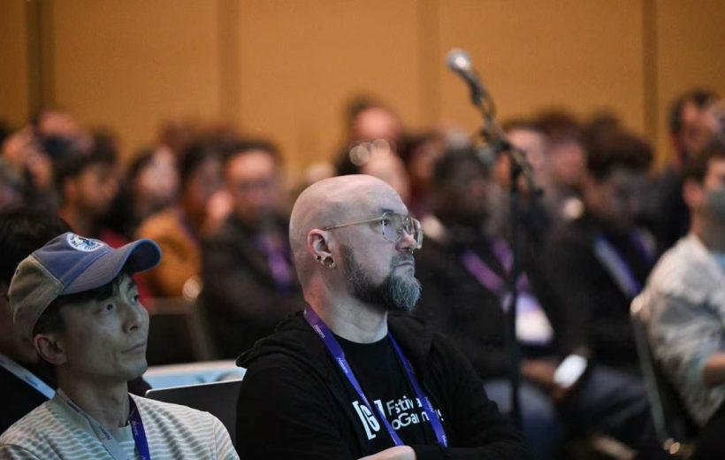 Bald man with glasses and lanyard attentively listening at a crowded conference session
