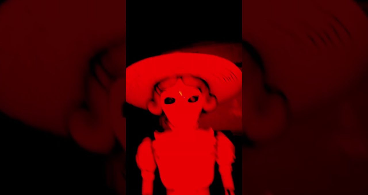 Red-tinted eerie doll wearing a wide-brimmed hat against a dark background