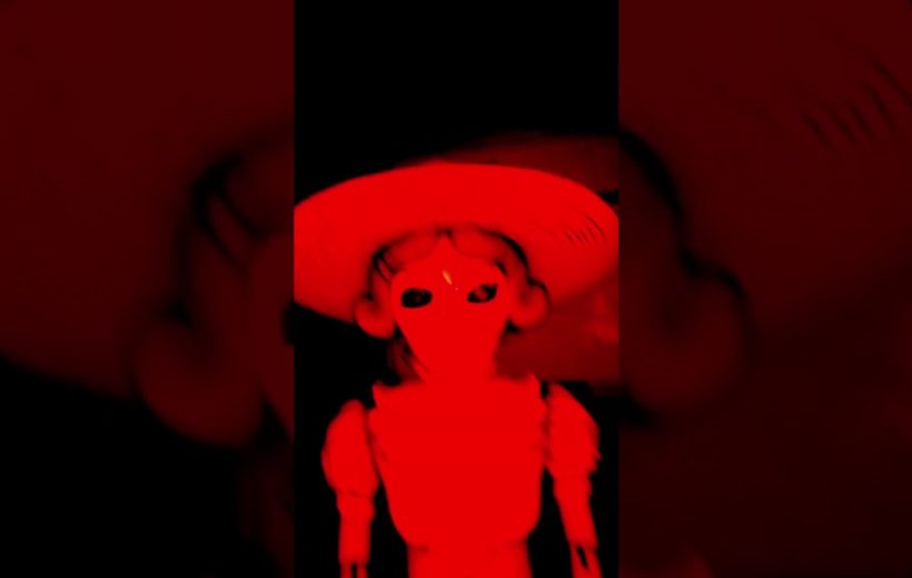 Red-tinted eerie doll wearing a wide-brimmed hat against a dark background