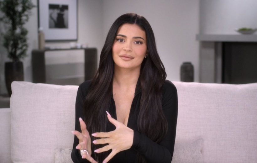 Woman with long dark hair sitting on a sofa, speaking and gesturing with hands