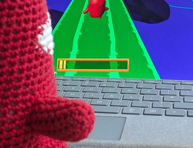 Crocheted red toy watching a colorful obstacle-run game on a laptop screen