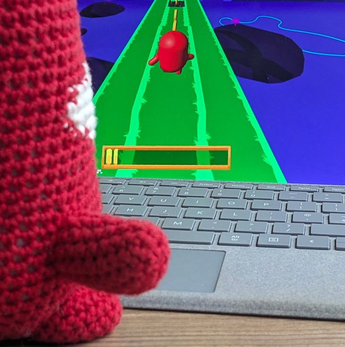 Crocheted red toy watching a colorful obstacle-run game on a laptop screen